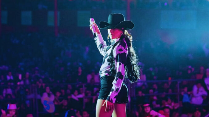 Ana Castela se apresenta em show de agronejo, usando chapéu e camisa com estampa rural, diante de um público iluminado por luzes de palco.