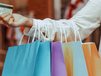 Pessoa segurando sacolas coloridas e cartão de crédito, representando compras por impulso estimuladas pelo consumo rápido e publicidade.