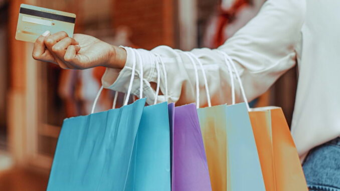 Pessoa segurando sacolas coloridas e cartão de crédito, representando compras por impulso estimuladas pelo consumo rápido e publicidade.